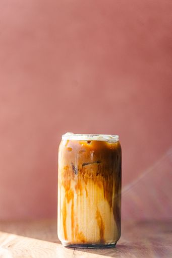 A refreshing iced coffee in a glass can with caramel swirls against a soft pink background, perfect for a summer day.
