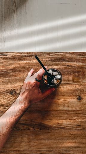 A vertical shot of a person holding a glass of iced coffee with a straw on the table