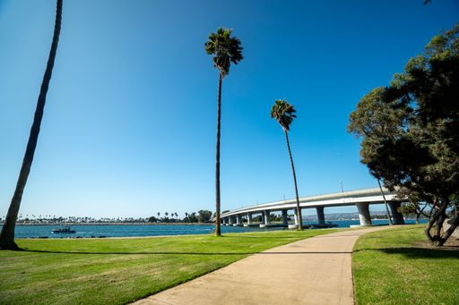 A concrete path through a park with a bridge in the background on a clear sunny day