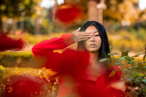 A portrait of an East Asian woman wearing a red suit and covering her eye with a hand in a garden