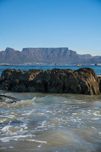 Scenic view of Table Mountain from the rocky shore of Cape Town with clear blue skies and gentle ocean waves.