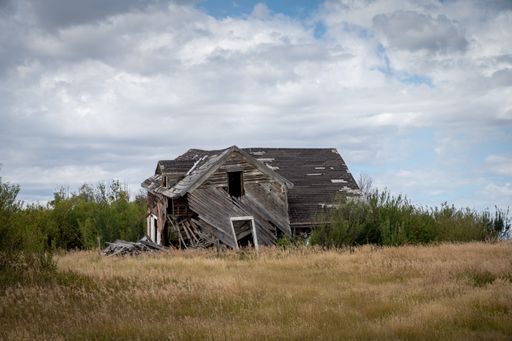 A horizontal shot of an old destroyed wooden home on a straw sunny field under a sky on the horizon
