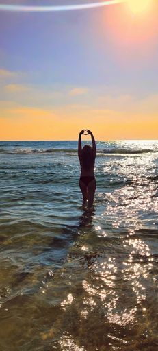 Silhouette of a woman in swimwear standing in the ocean at sunset, forming a heart shape with her hands against the vibrant sky.