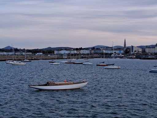 A serene view of sailboats anchored in Dun Laoghaire harbor with a backdrop of coastal town buildings and distant hills under a cloudy sky.
