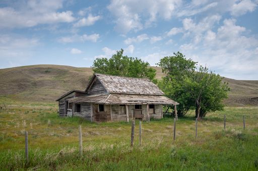 A horizontal shot of an old destroyed wooden home on a sunny green field under a sky on the horizon
