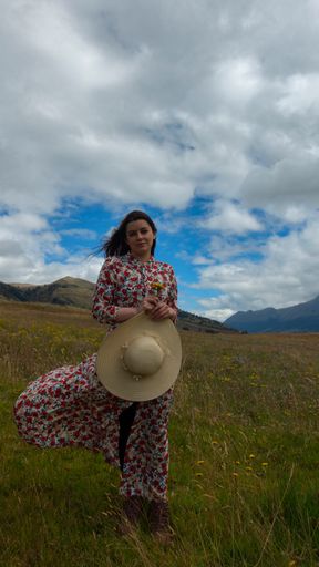 A happy young Hispanic woman in a floral dress and hat walking in a wheat field on a cloudy day