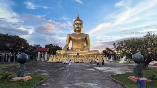 A large golden Buddha statue in a Thai temple complex under a vibrant blue sky with scattered clouds, people of various ethnicities around.
