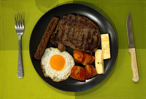 Grilled steak, sausage, egg, tomatoes, and halloumi served on a black plate with utensils.