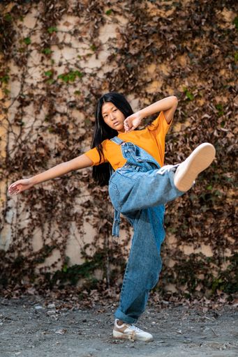 A pretty East Asian lady with dark hair and jeans overalls posing while demonstrating dance moved