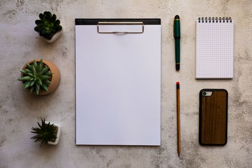 A flat lay shot of a clipboard, a notebook, pens and houseplants on a desk under the sunlight