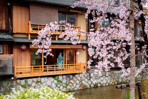 A traditional Japanese house with cherry blossoms in full bloom, featuring a woman in a kimono standing on the balcony.