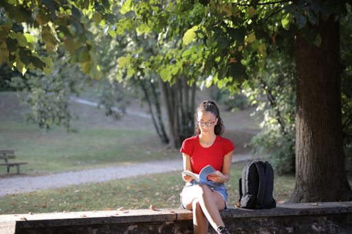 A young girl with glasses sitting under the tree in the park and reading a book