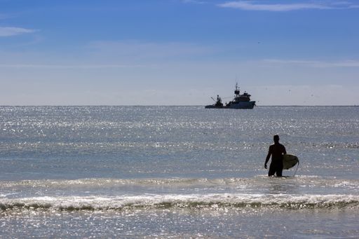 A shot of a surfer walking in the sea with a ship in the background