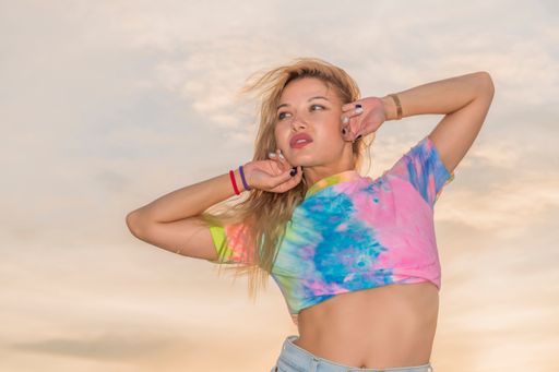 Young tanned girl in bright sportswear stands in background of bright orange summer sky. Looks into distance, raised hands and brought behind her head