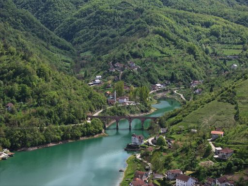 Aerial view of a picturesque village nestled in lush green mountains with a serene river and a charming bridge.