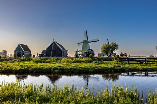 Scenic view of traditional Dutch windmills and houses with clear blue sky in Zaanse Schans, Netherlands, reflected in a calm canal.