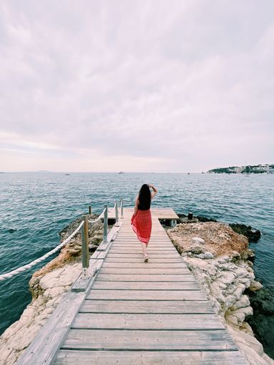 A vertical shot of a woman in a red skirt walking on a jetty stretching out to the sea under a cloudy