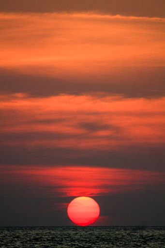 A vertical shot of the round sun setting down on the horizon by the ocean with breathtaking sky