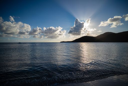 A scenic view of a tranquil beach at sunset with sun rays peeking through clouds over a calm sea