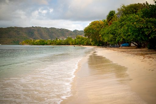 A serene tropical beach with gentle waves, lush greenery, and distant hills under a cloudy sky.