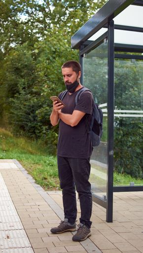 A vertical shot of a Hispanic guy standing at the bus stop and looking at his phone