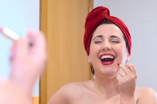A young female with a red towel on her head applying red lipstick while standing in front of a mirror in the bathroom