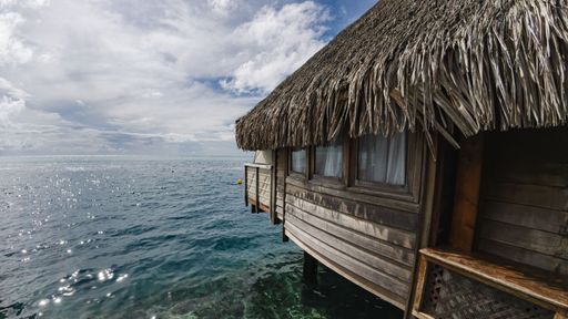 A serene overwater bungalow with a thatched roof overlooks the sparkling ocean under a partly cloudy sky, evoking tropical paradise vibes.