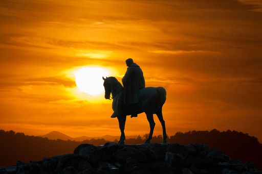 Silhouette of a statue of a rider on horseback against a vibrant sunset sky, creating a dramatic and historical scene.