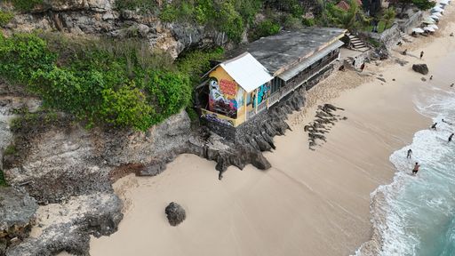 Aerial view of a beachside building with vibrant graffiti, surrounded by cliffs and sandy shore, with people enjoying the water in Bali, Indonesia.