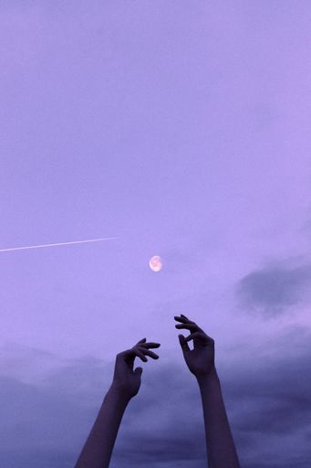 A closeup of hands reaching to the crescent of the moon on a night sky