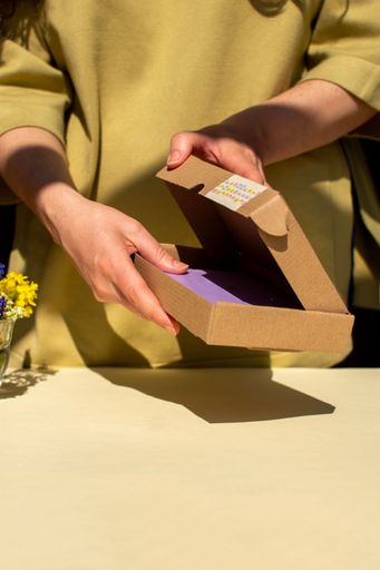 A person opening a cardboard box with a colorful label on a sunny day, with a bouquet of flowers on the table.