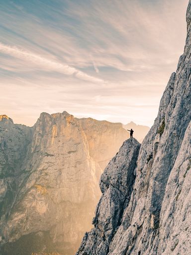 Hiker or climber standing on high rock pointing towards sunrise. Glowing mountain landscape with a silhouette in the glowing sun. Adventure ascent.