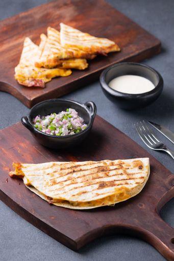 A vertical shot of a quesadilla on a wooden board with a bowl of chopped onion and jalapeno