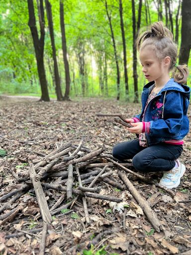 A curious young girl kneels in a sun-dappled forest, constructing a small stick teepee from twigs and branches scattered on the leaf-strewn ground