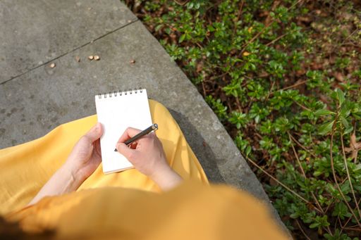 A person in a yellow dress writes in a notebook while sitting outdoors, surrounded by greenery.