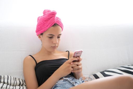 A Caucasian young woman with a pink towel on her head sitting on a bed while using a mobile phone