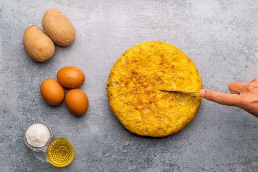 Spanish omelette with ingredients on the side on concrete texture background. Hand cutting a portion of the tortilla. Overhead view.