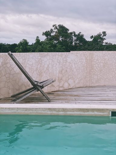 Minimalist poolside scene with a wooden lounge chair and cloudy sky, evoking a tranquil atmosphere.