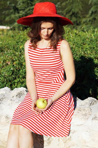 A young Caucasian female in a striped dress and a red hat holding an apple