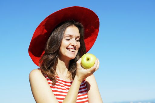 A young Caucasian female in a striped dress and a red hat eating an apple