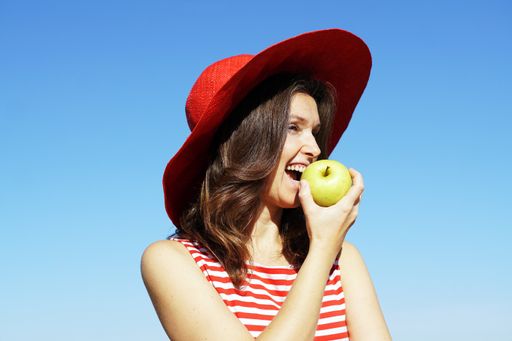 A young Caucasian female in a striped dress and a red hat eating an apple