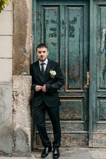 Full length portrait of young groom wearing black suit standing in front of old green door.