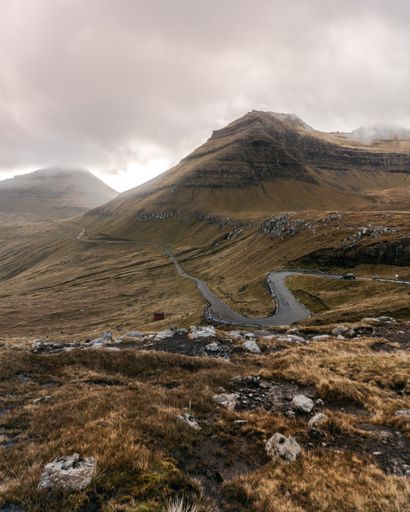 S-curved road leads to the mountains through the hills under cloudy sky. Moody Scandinavian landscape. Visit Faroe Islands