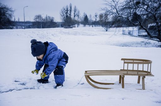 A little cute boy in winterwear playing with snow next to a wooden sled at a park on a cold winter day