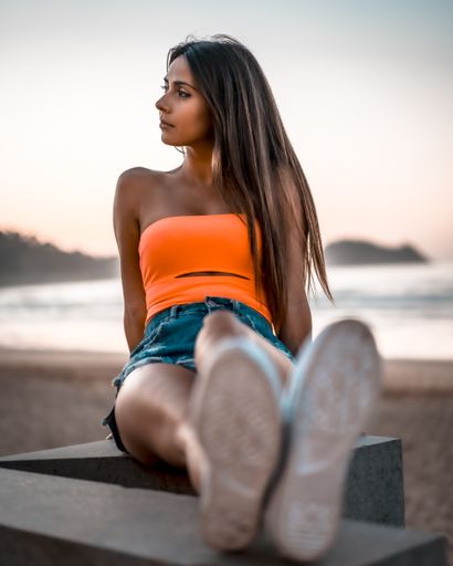 A vertical shot of a young brunette female in jeans shorts and an orange top sitting in the beach of Zarautz, Spain in summer