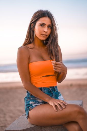 A vertical shot of a young brunette female in jeans shorts and an orange top sitting in the beach of Zarautz, Spain in summer