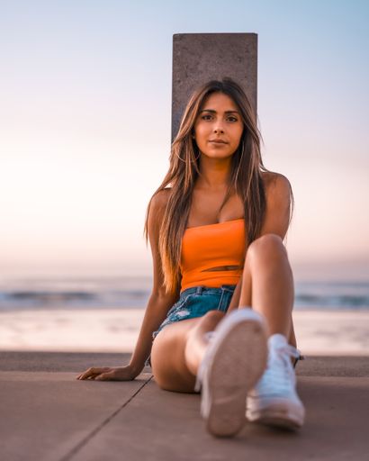A vertical shot of a young brunette female in jeans shorts and an orange top sitting in the beach of Zarautz, Spain in summer