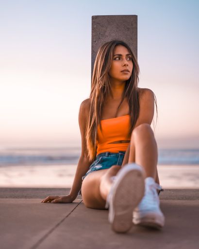 A vertical shot of a young brunette female in jeans shorts and an orange top sitting in the beach of Zarautz, Spain in summer