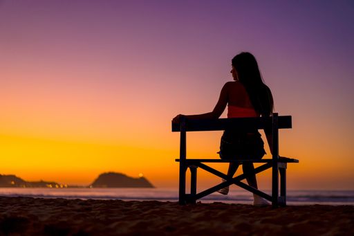 A female sitting on a bench in the beach with the colorful sky in the background
