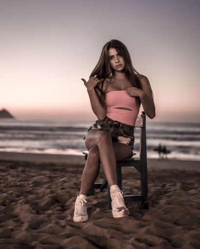 A vertical shot of an attractive brunette sitting on a wooden chair with legs crossed at the beach in Zarautz, Spain